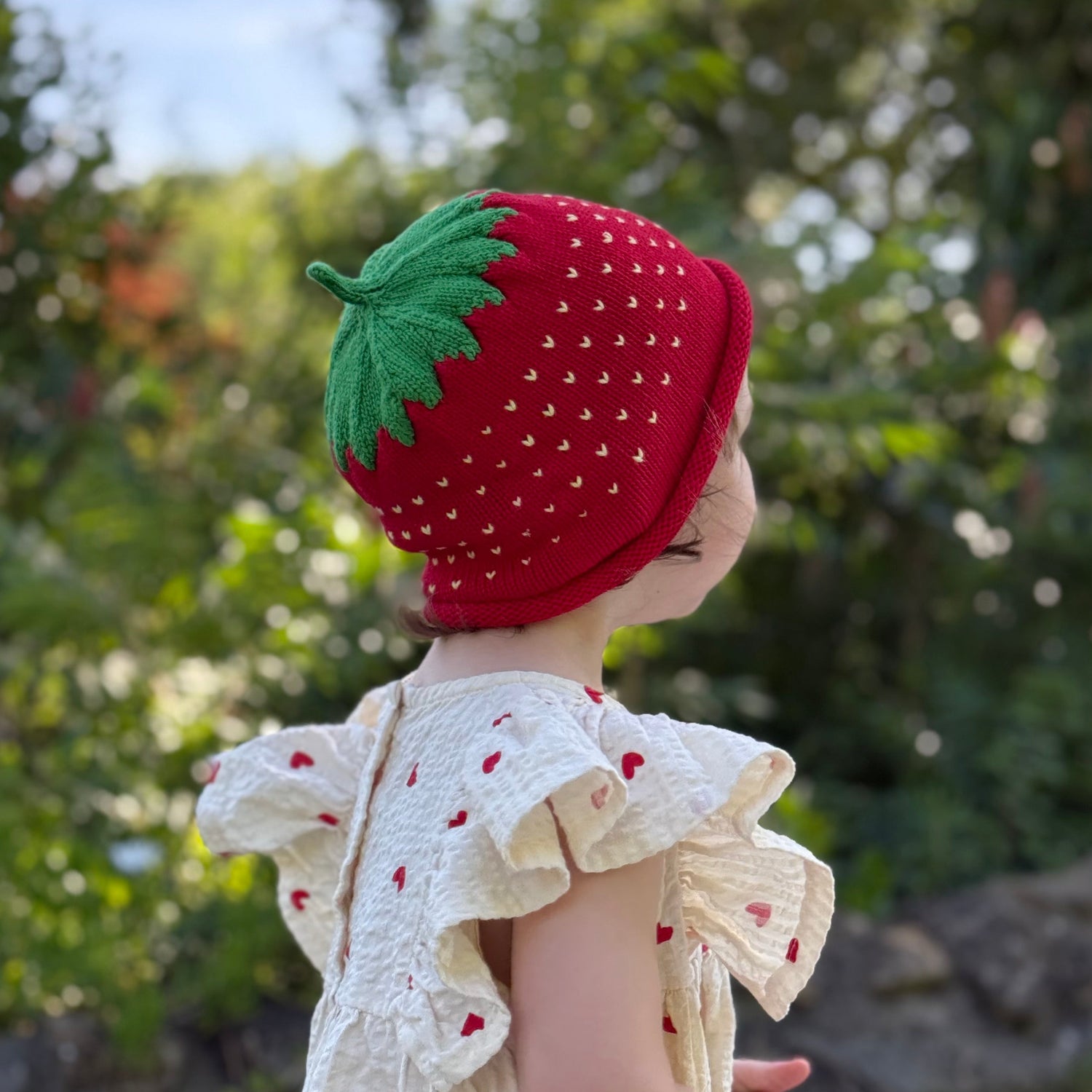 Child wearing a strawberry-themed hat outdoors with greenery in the background