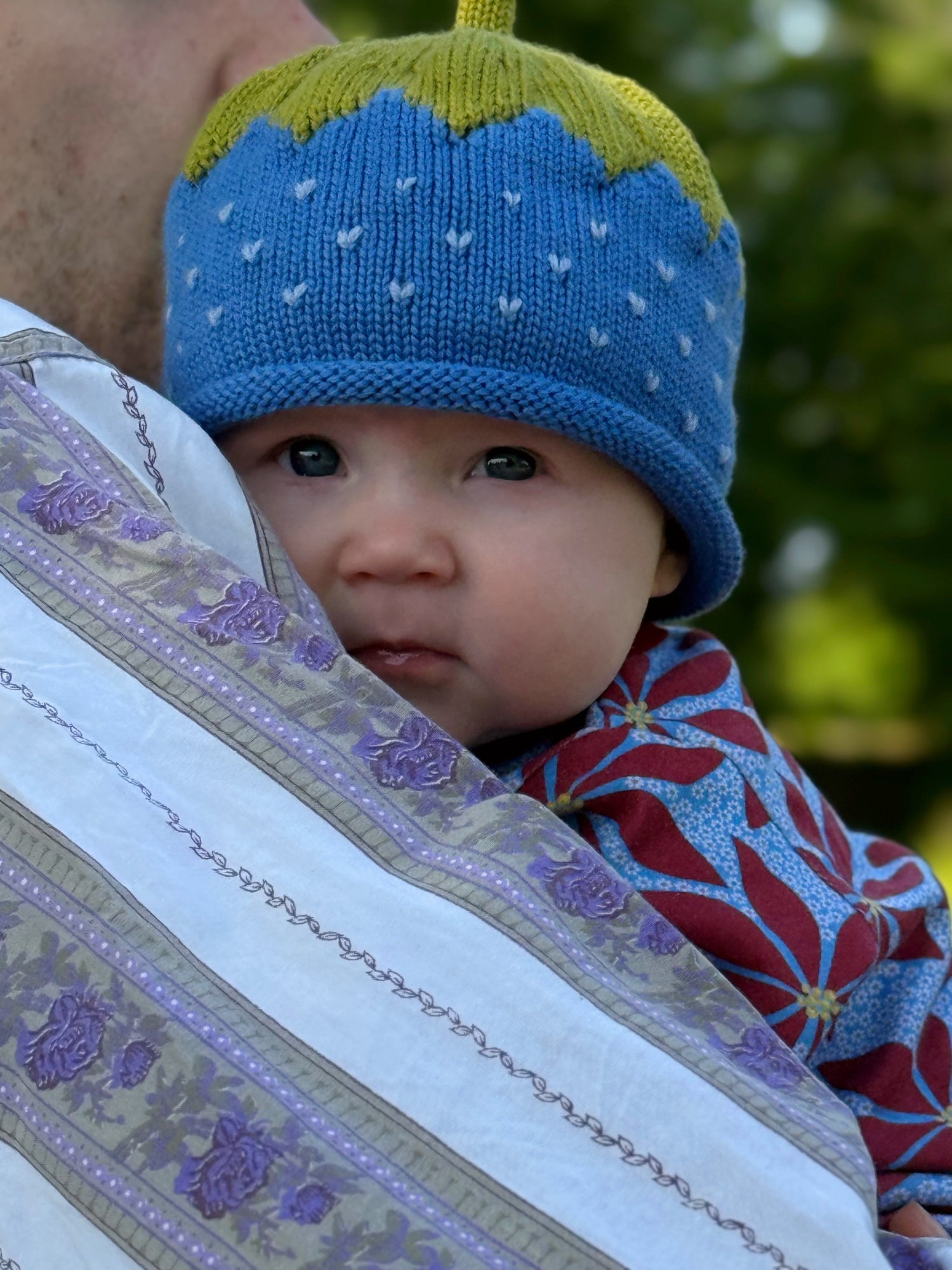 Baby wearing a blue and green knit hat with white dots, peeking out from behind an embroidered fabric.