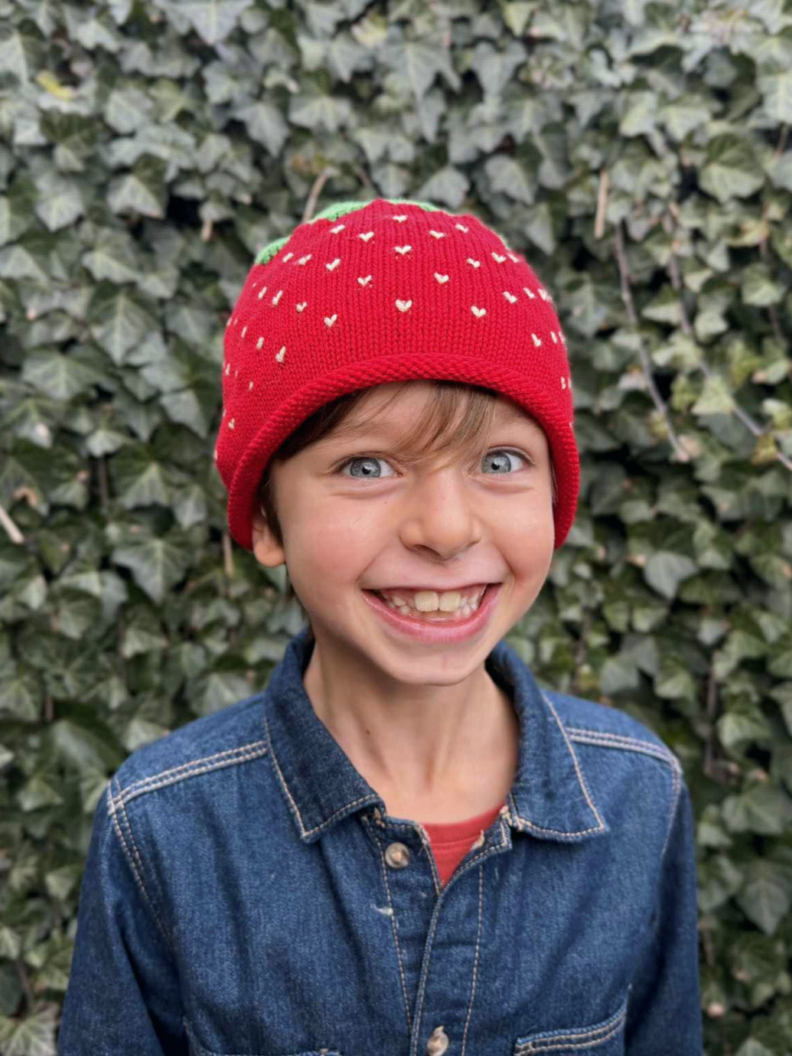 Child wearing a red knit hat with white patterns against a green ivy background