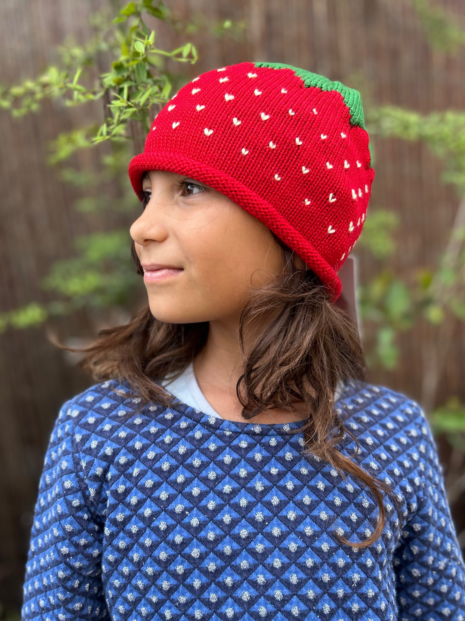 Child wearing a red knit strawberry hat with green leaves against a blurred natural background