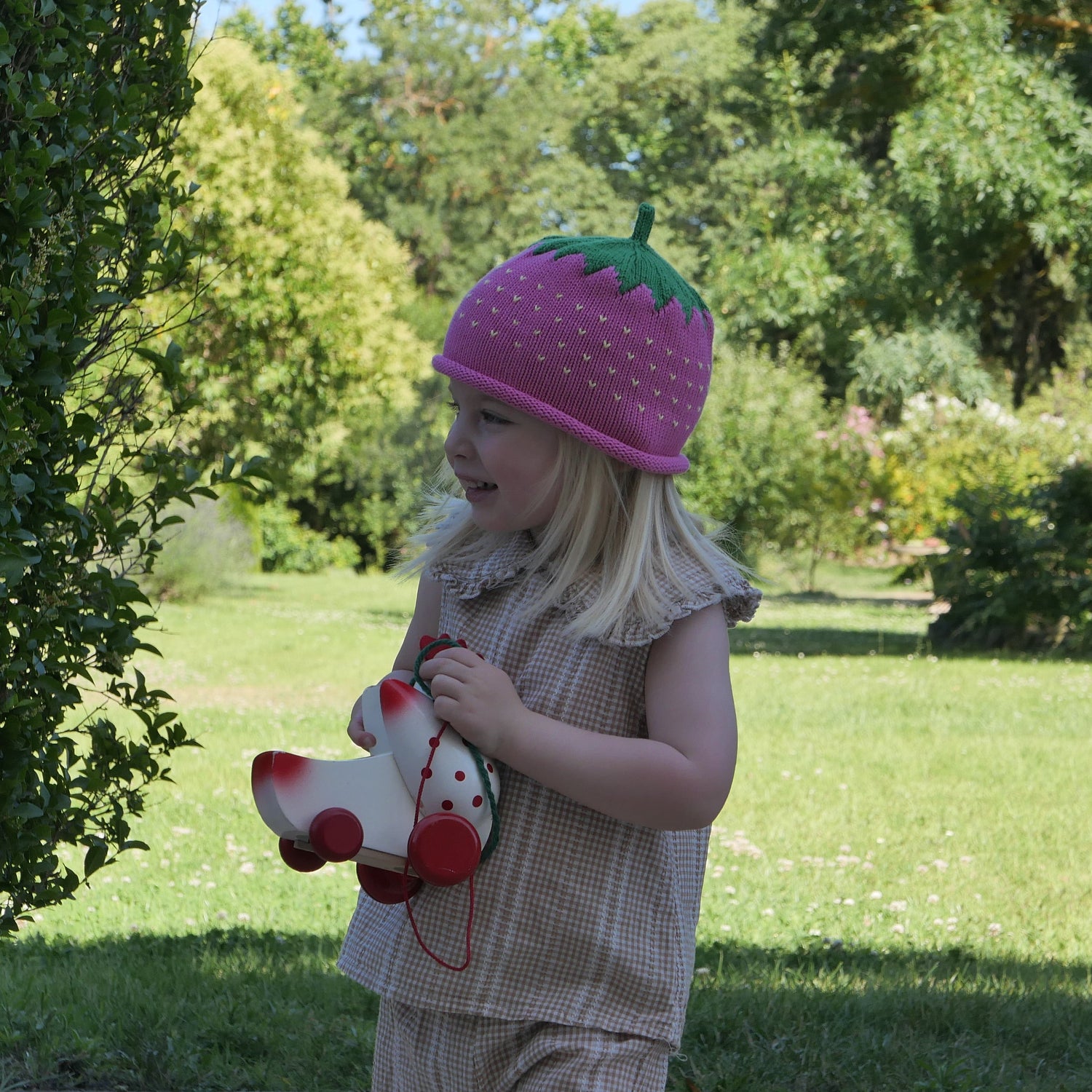 Child wearing a pink knit hat with a green stem and red berries, standing outdoors in a garden.