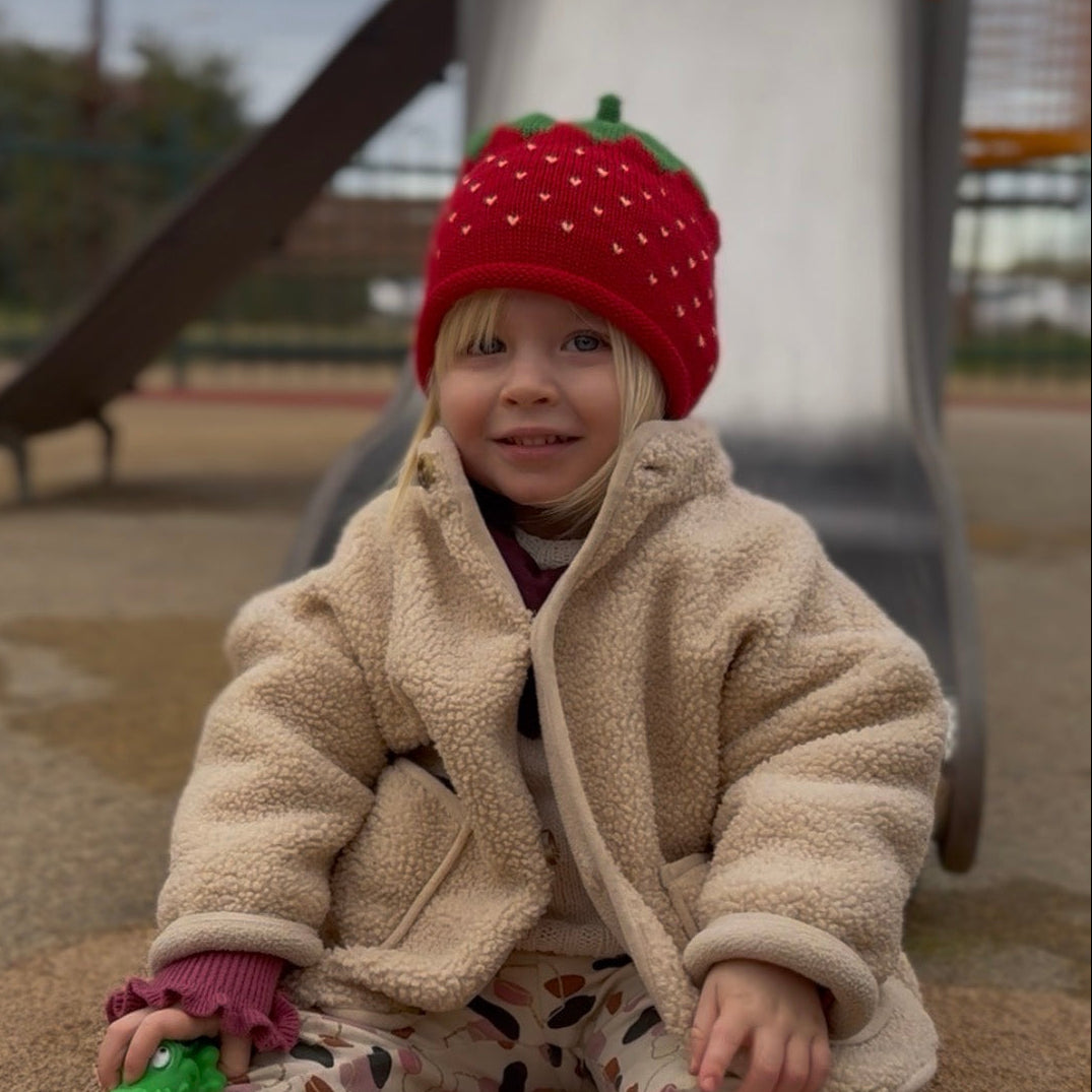 Child wearing a red strawberry hat and beige coat sitting on a playground.