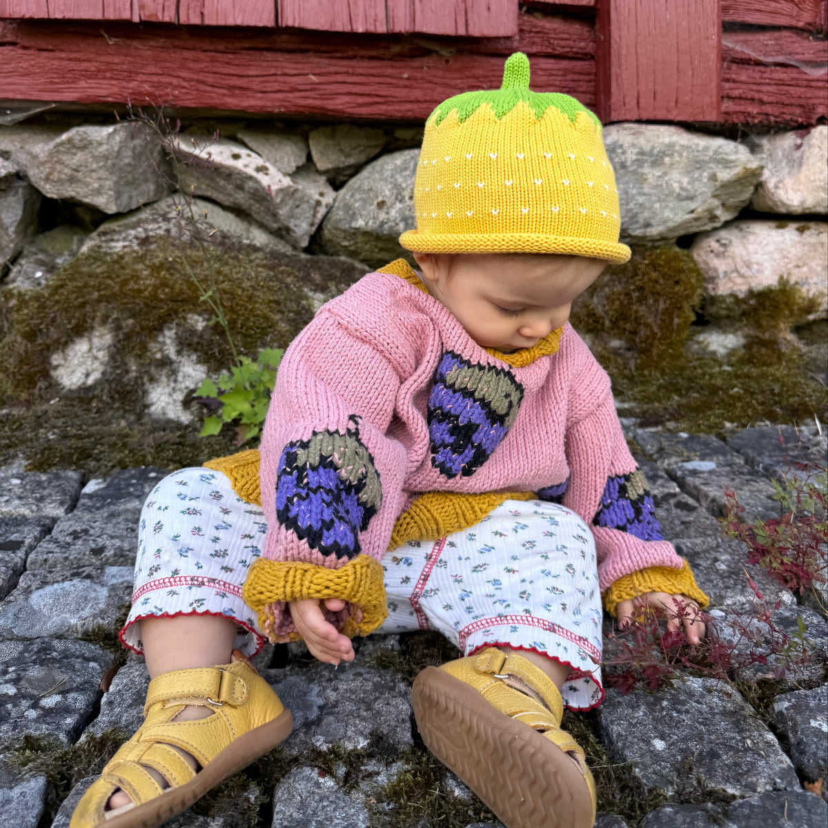 A baby sitting outdoors on a stone surface in front of a red wooden wall, wearing a pink knit sweater with purple and yellow strawberry motifs, a yellow strawberry-shaped hat, patterned pants, and yellow sandals.