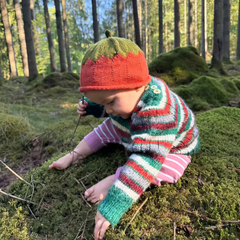 A toddler kneeling on moss in a forest, wearing a colorful knit pumpkin hat with a red top and green bottom, a striped knit sweater, and pink leggings.