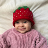 Baby wearing a pink knitted sweater and a red strawberry hat on a white background
