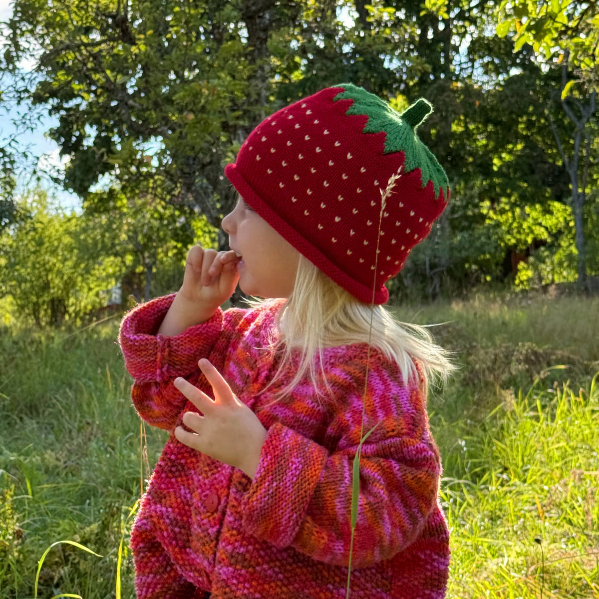 A young girl wearing a red strawberry-themed knit beanie and a pink and red knit sweater stands in a grassy outdoor field with trees in the background.