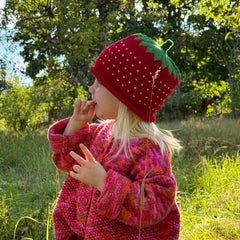 A young girl wearing a red strawberry-themed knit beanie and a pink and red knit sweater stands in a grassy outdoor field with trees in the background.