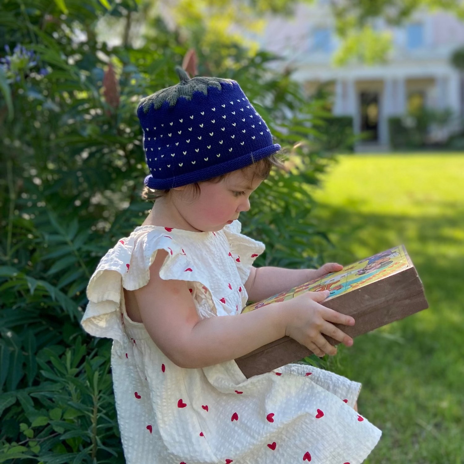 Child in a white dress with red hearts, blue polka dot hat, and brown book sitting on grass.