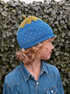 Person wearing a blue knit hat with a yellow peak against a green hedge background