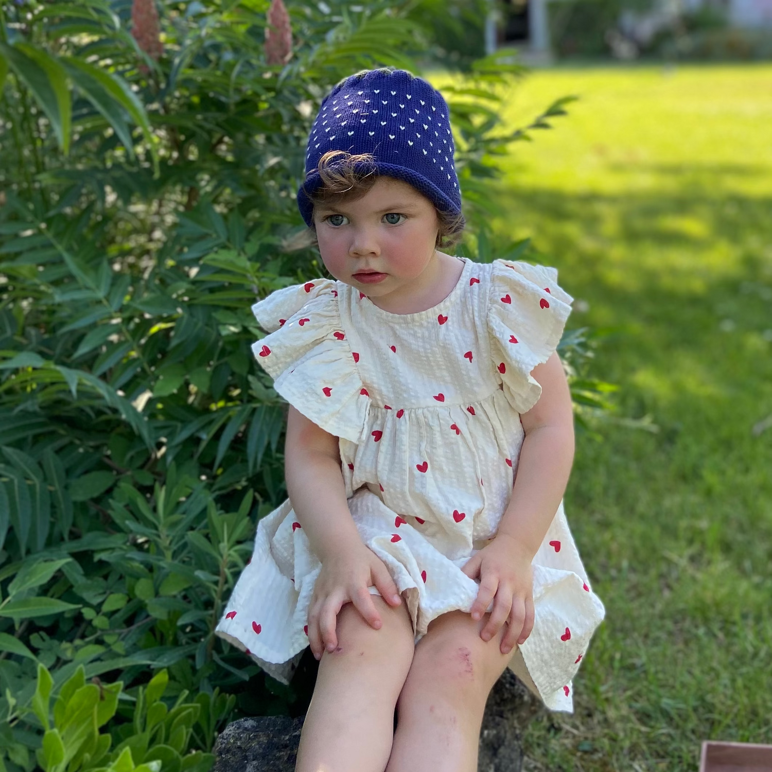 Child in a white dress with red polka dots and blue hat sitting on grass with a house in the background