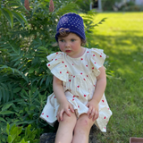 Child in a white dress with red polka dots and blue hat sitting on grass with a house in the background