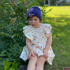 Child in a white dress with red polka dots and blue hat sitting on grass with a house in the background