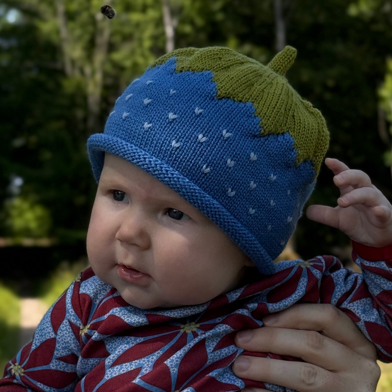 Baby wearing a blue and green knit hat with a leaf design, held by an adult outdoors.