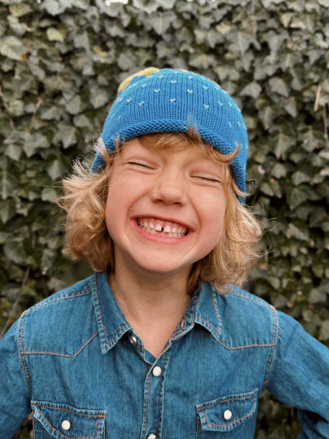 Child wearing a blue knit hat and denim shirt against a green leafy background