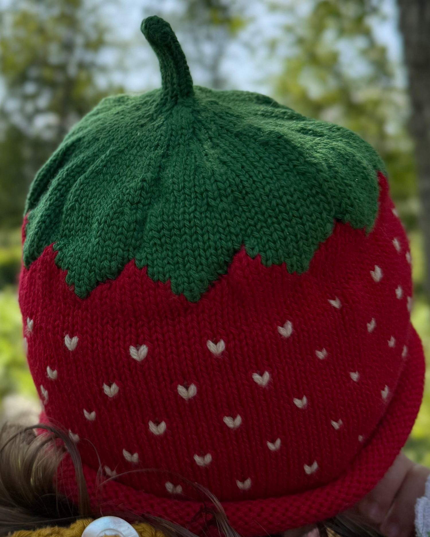 Strawberry-shaped knitted hat with green top and red body with white spots, worn outdoors.