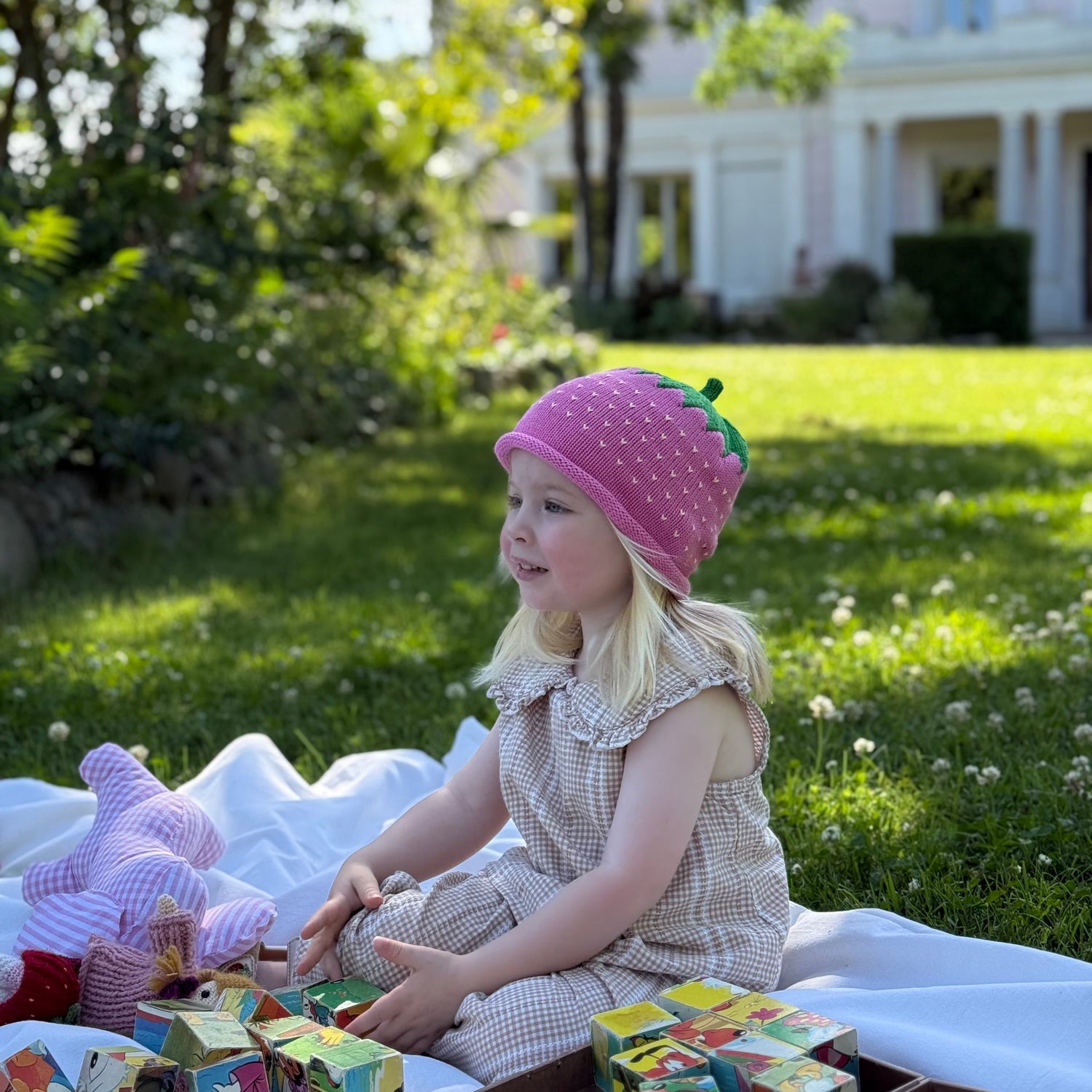 Child sitting on a blanket in a garden with a pink hat and colorful blocks.