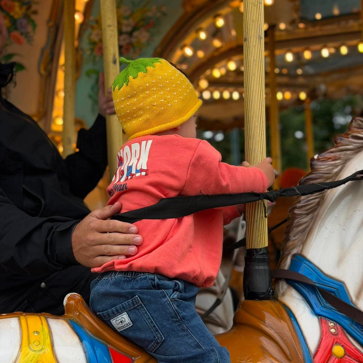 A toddler wearing a bright yellow lemon hat and a coral pink sweatshirt sits on a carousel horse, holding onto a wooden pole. An adult in a black jacket supports the child from behind.