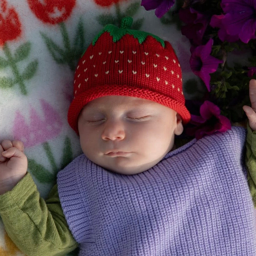 A sleeping newborn baby wearing a red strawberry-shaped knit hat and a light purple blanket, lying on a white blanket with red and green floral patterns next to purple flowers.