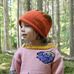 A young child wearing a knitted orange pumpkin hat with green leaves and a pink sweater with a blue and green pumpkin motif, standing in a forest.