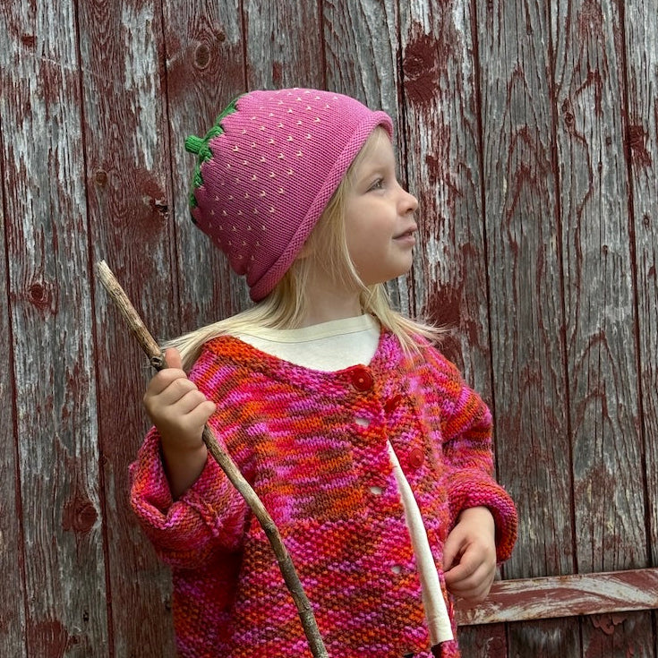 A young child wearing a pink strawberry-themed knit hat and a vibrant pink and orange patterned cardigan, holding a wooden stick in front of a weathered red-brown wooden barn door.