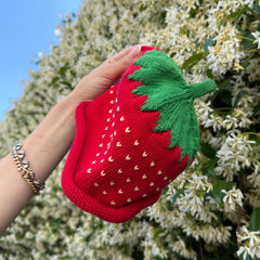 Hand holding a red knitted strawberry hat in front of a flowering bush