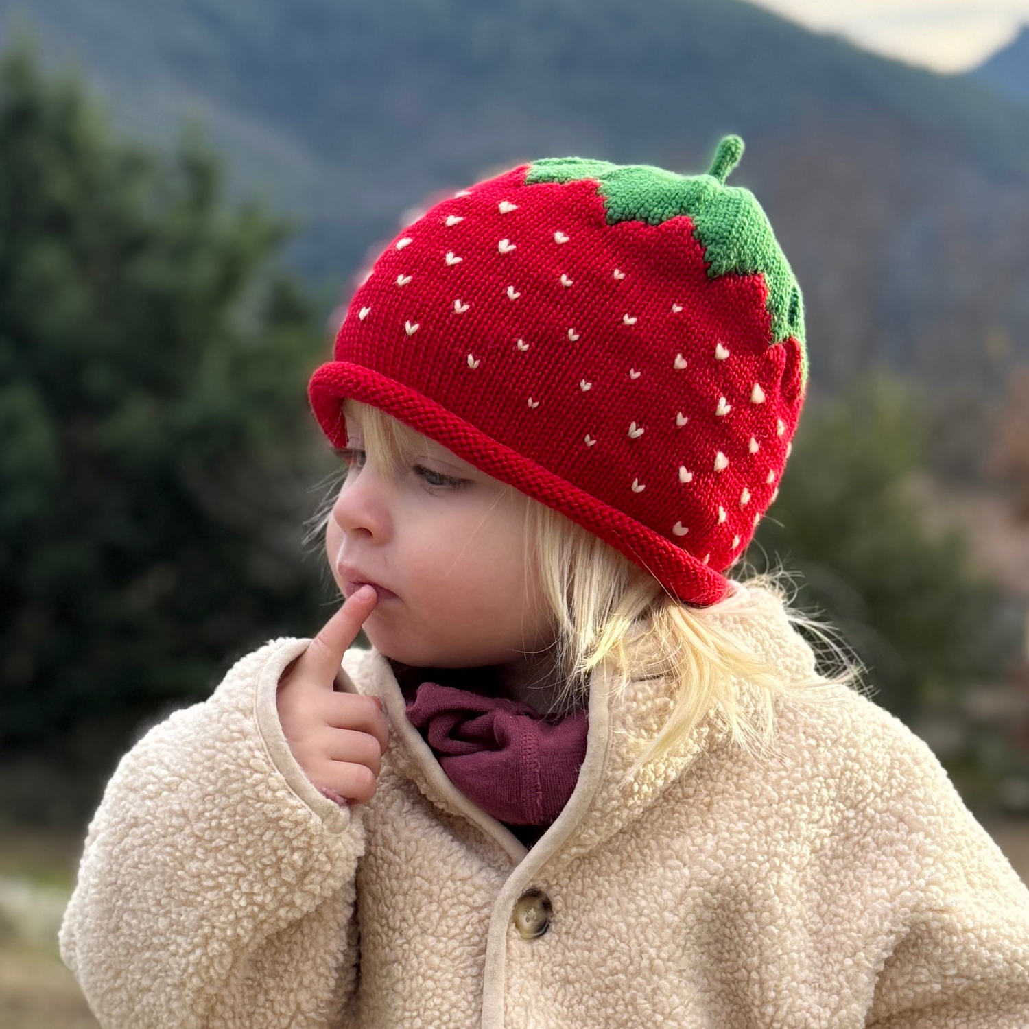 Child wearing a red strawberry hat outdoors with mountains in the background
