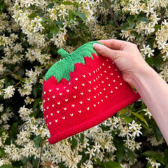 Hand holding a red strawberry-shaped hat against a background of white flowers.