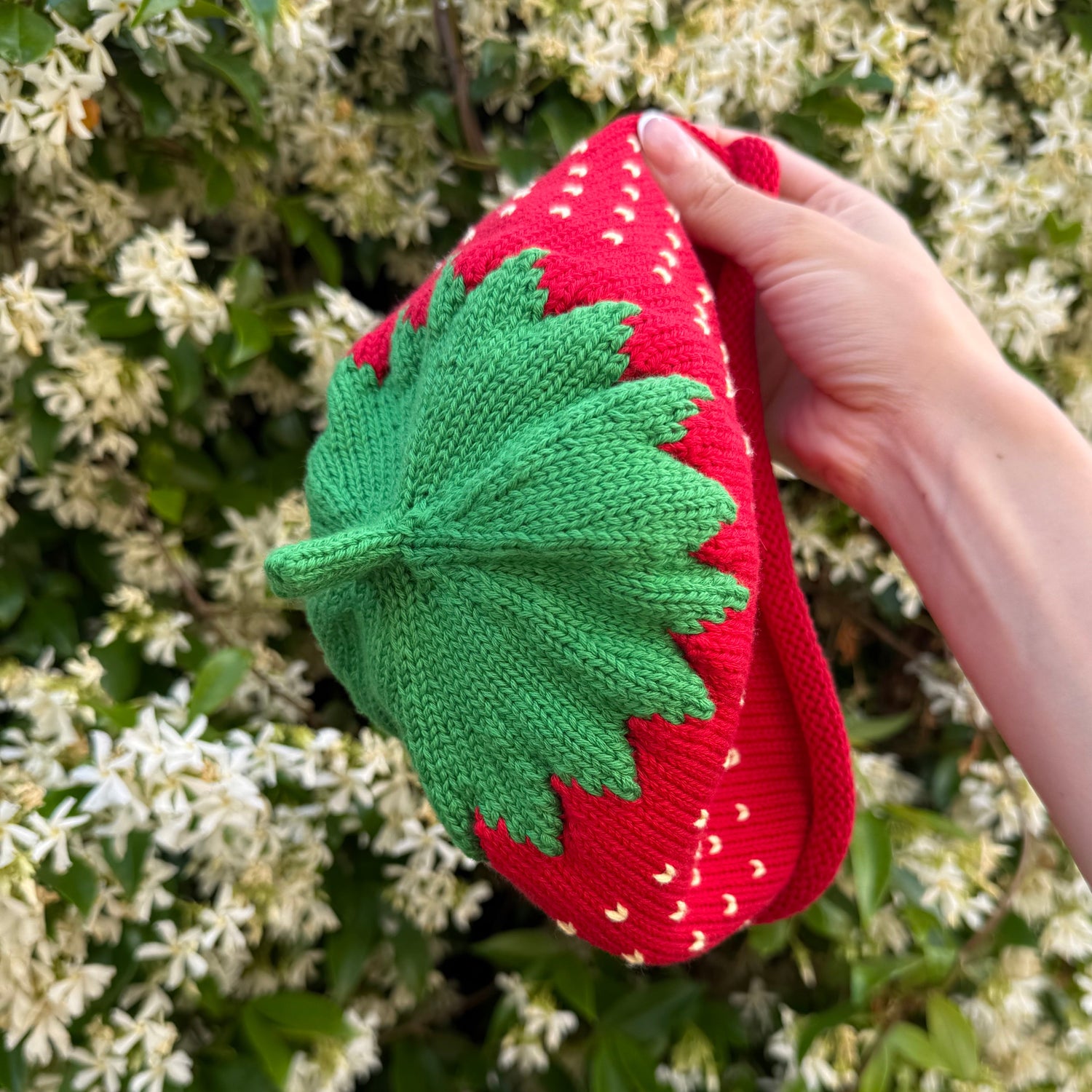 Hand holding a red and green knitted strawberry hat against a white flower background