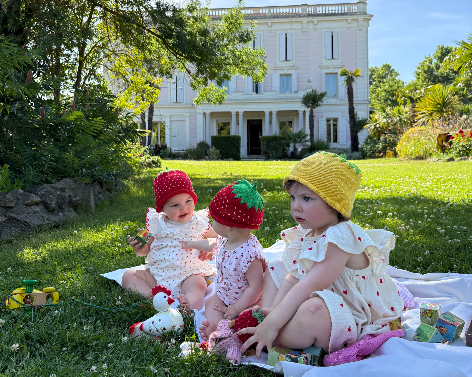 Three children in colorful strawberry hats sitting on a blanket in a garden with a large house in the background.